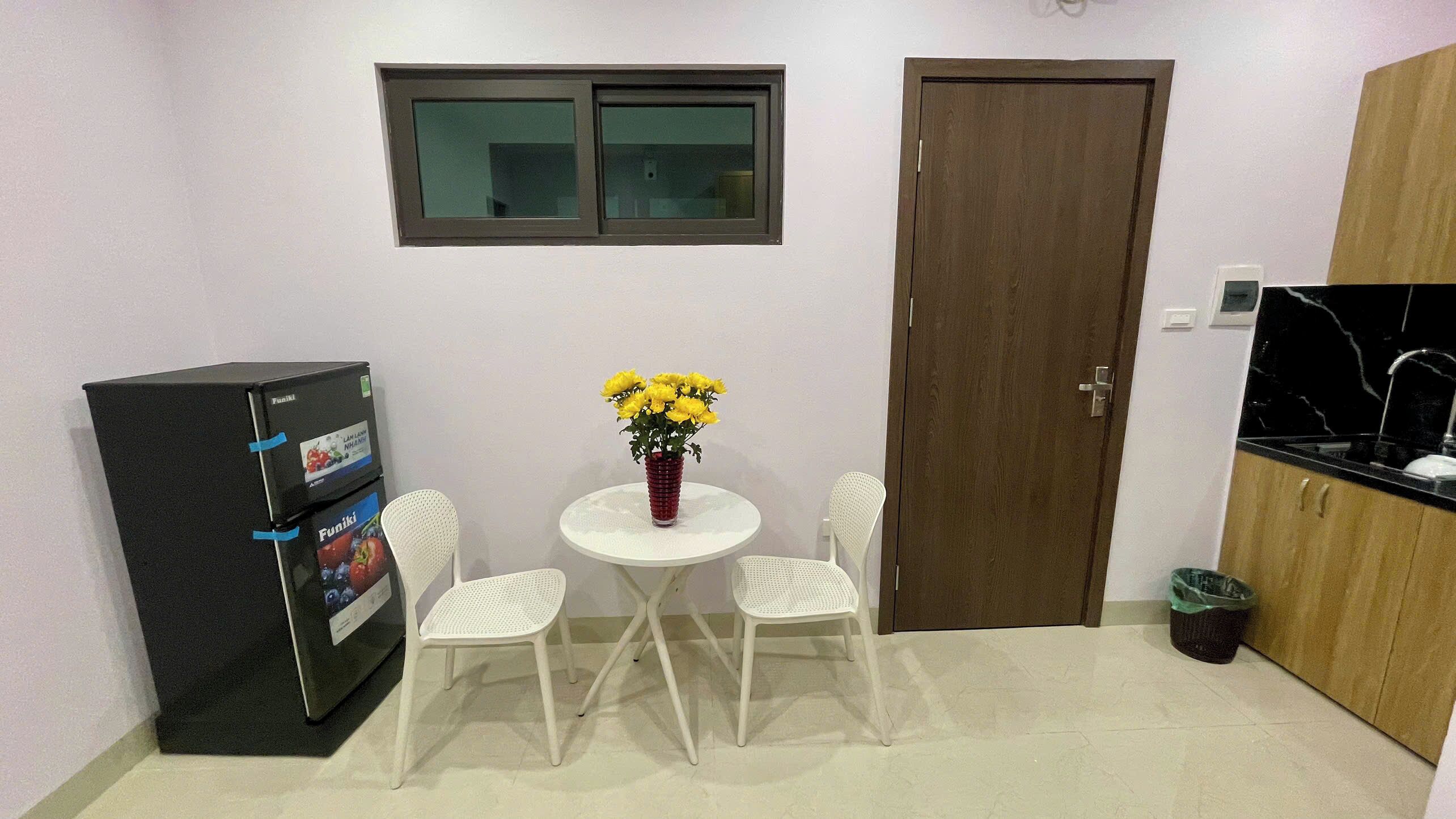 A small, clean room with a refrigerator, a table with chairs, a vase of flowers, and a door.