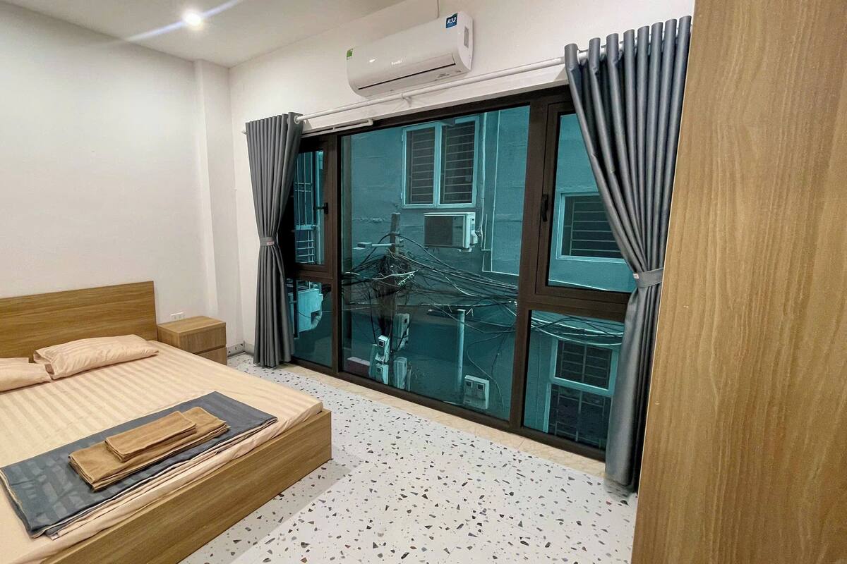 A bedroom with a bed, a nightstand, a window with curtains, and an air conditioning unit.