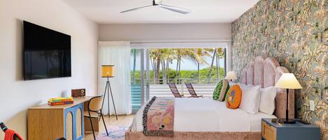 A bedroom with a bed, a desk, a chair, a TV, a ceiling fan, and a view of palm trees.