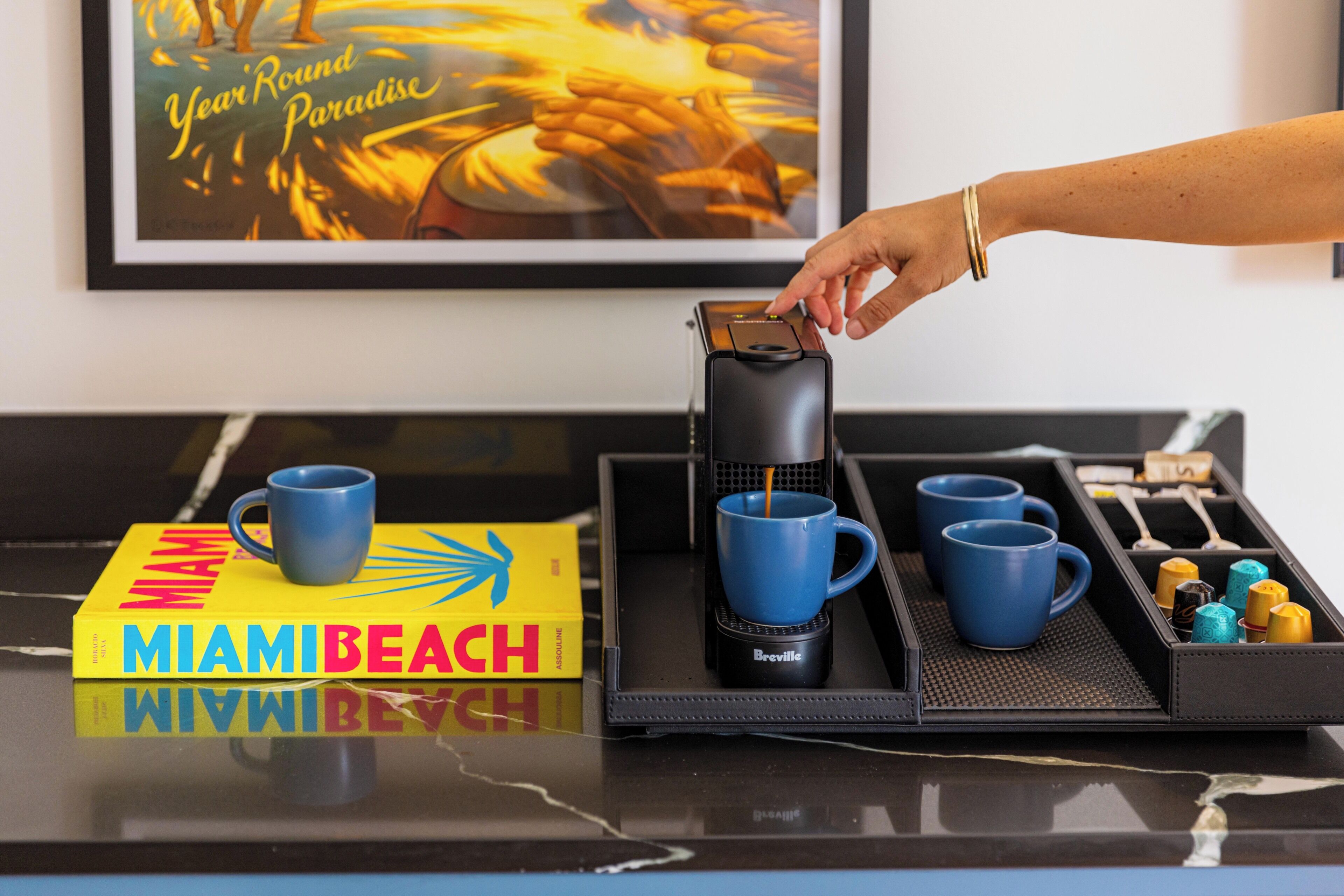 Ein Tablett mit einer Kaffeemaschine, blauen Tassen und einem Buch mit dem Titel 'MIAMI BEACH' auf einer spiegelnden Oberfläche.