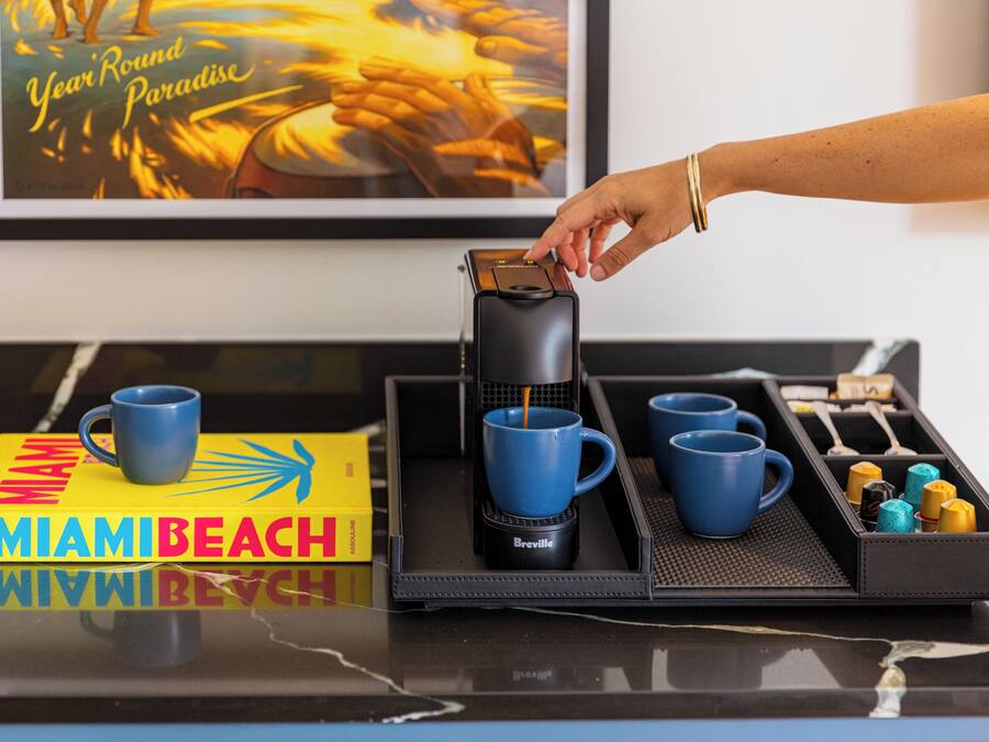 Ein Tablett mit einer Kaffeemaschine, blauen Tassen und einem Buch mit dem Titel 'MIAMI BEACH' auf einer spiegelnden Oberfläche.