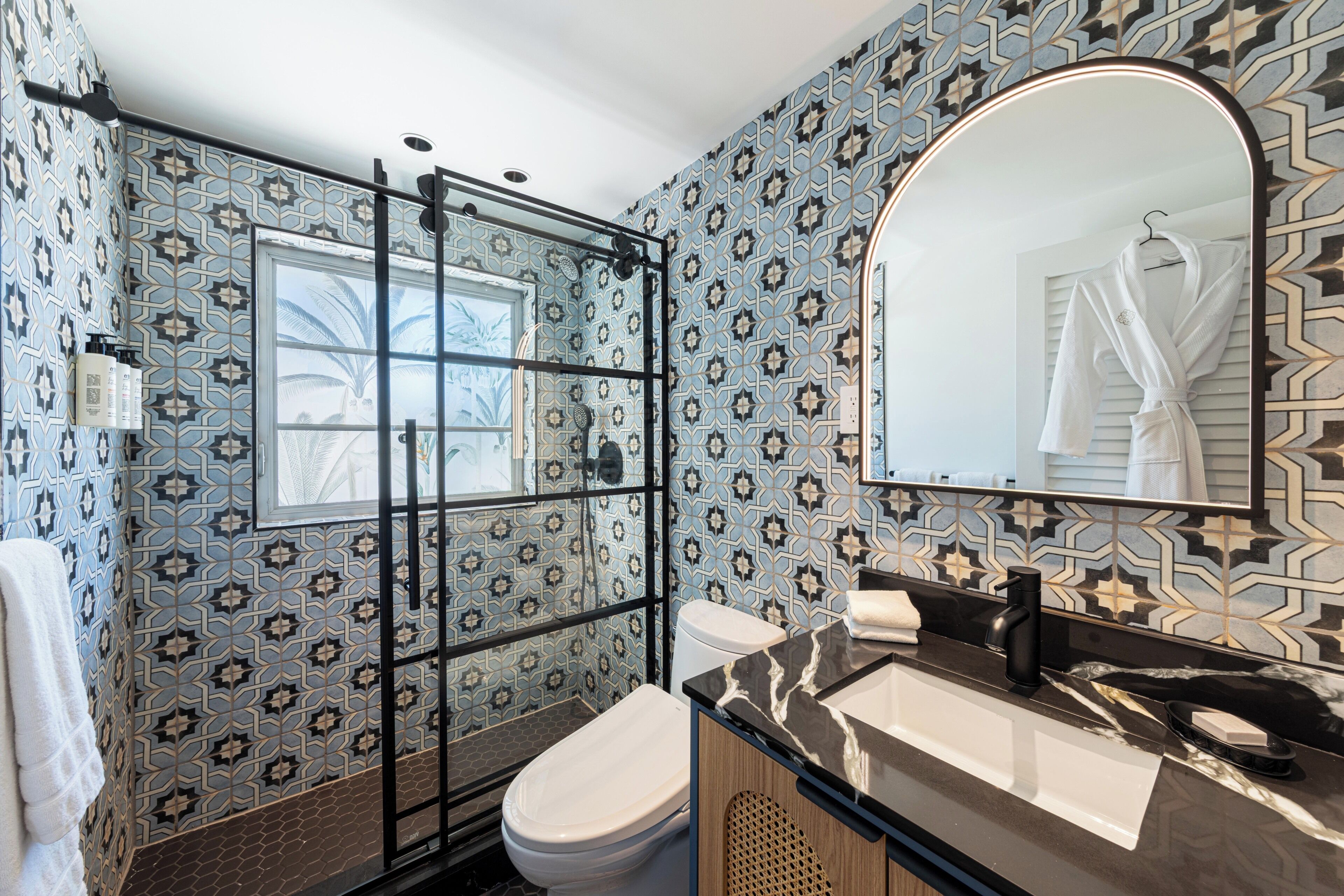 A bathroom with patterned tiles, a glass shower enclosure, a toilet, a sink with a marble countertop, and a mirror with a robe hanging.