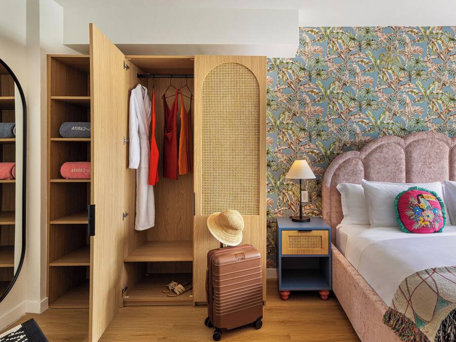 A hotel room with a wardrobe, a suitcase, a bed with pillows, a nightstand, and a wall with a floral pattern.