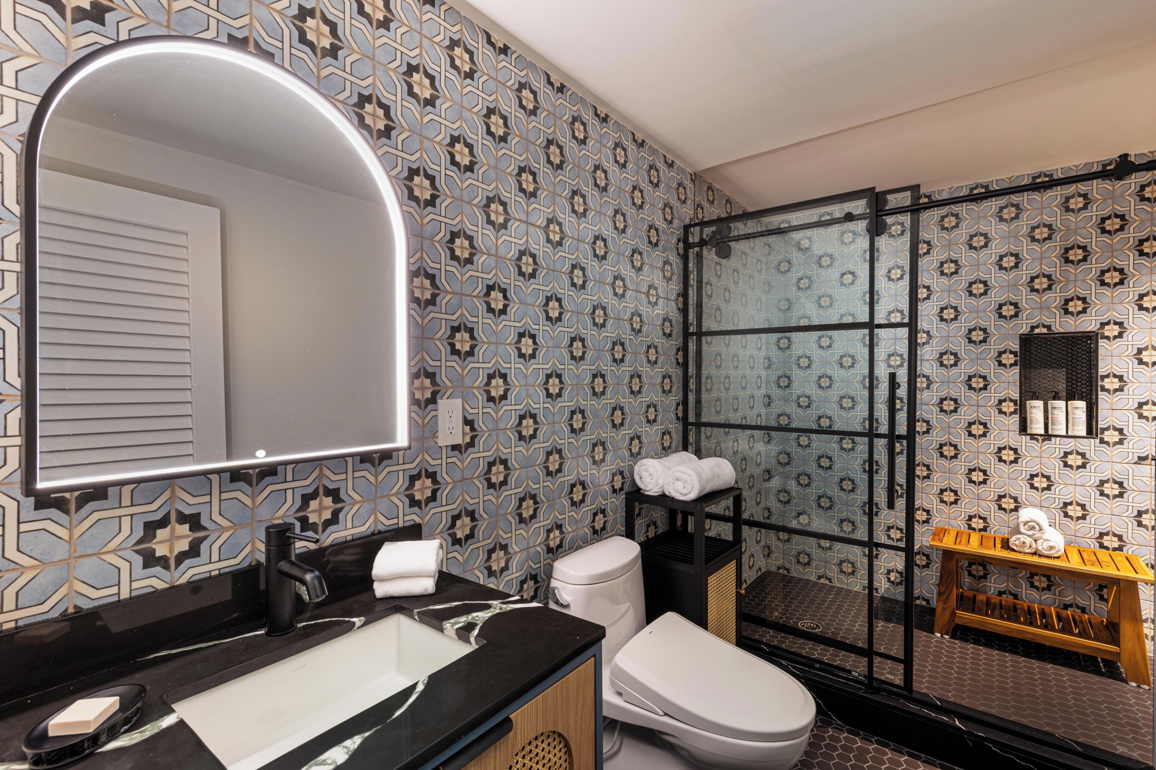 Un baño con pared de azulejos estampados, encimera negra, inodoro blanco, ducha con mampara de vidrio y estante de madera con toallas.