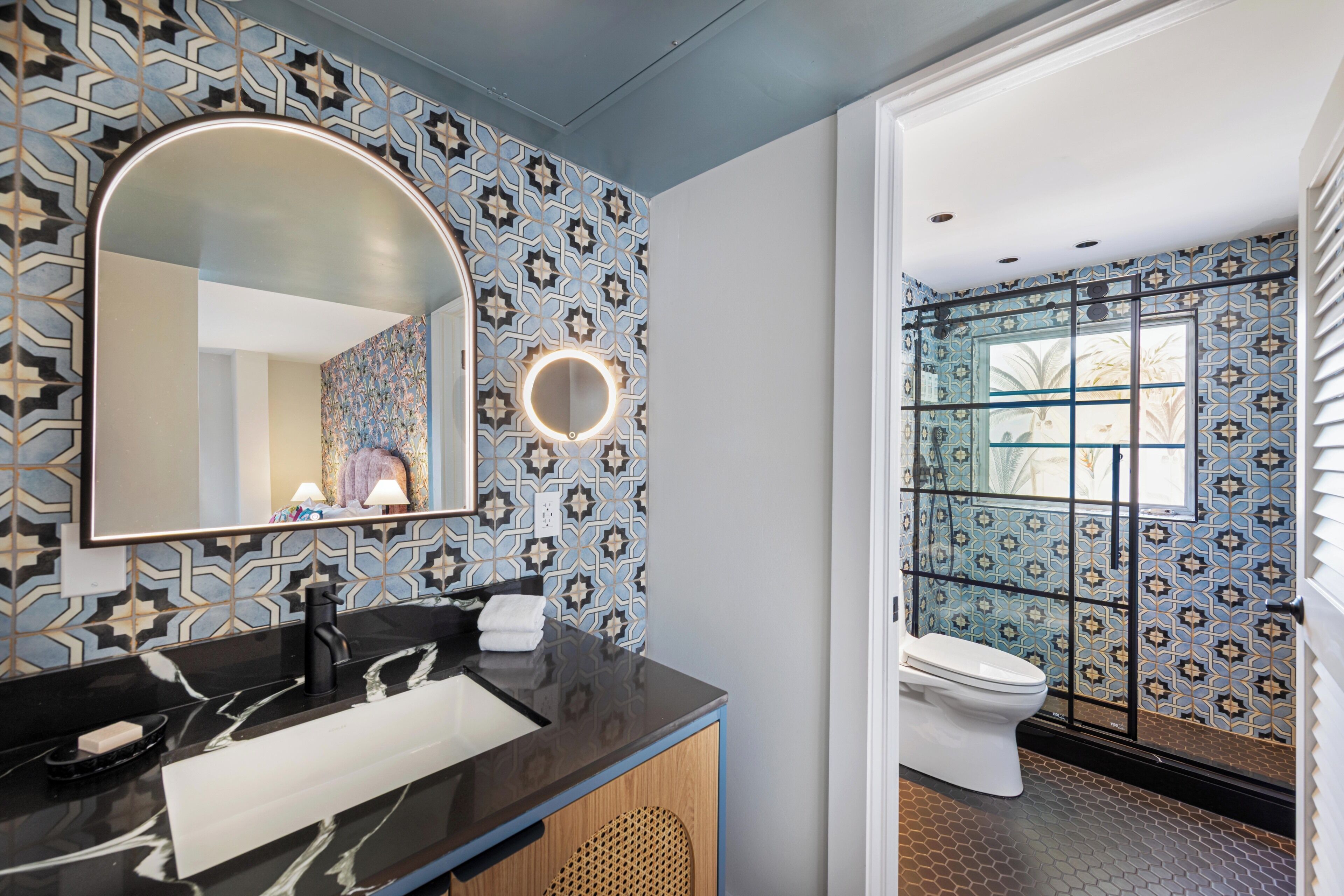 Une salle de bain avec un lavabo en marbre noir, un grand miroir avec un éclairage circulaire, des carreaux décorés et des toilettes avec une fenêtre.