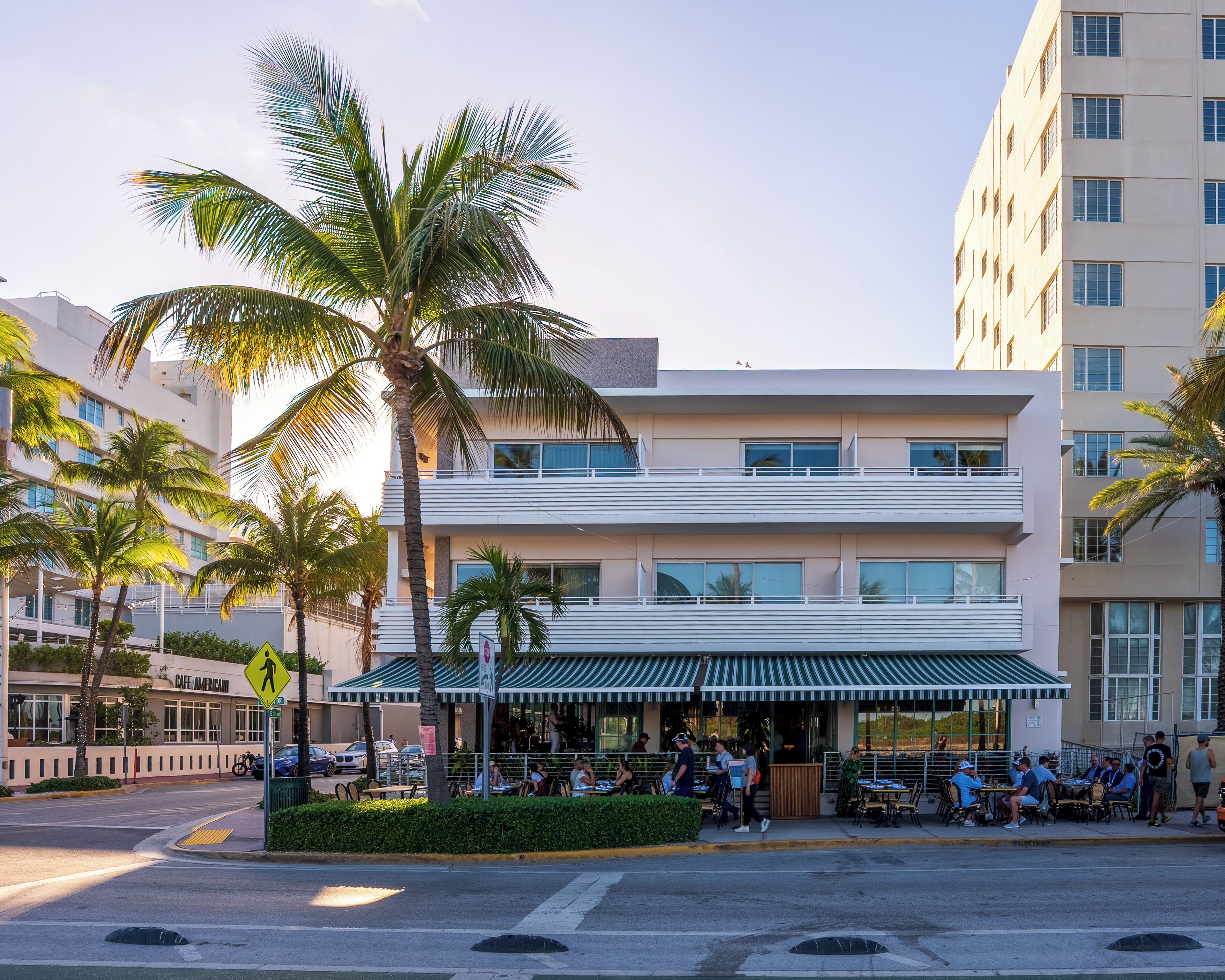 A modern building with a cafe patio, palm trees, and a pedestrian crossing sign.