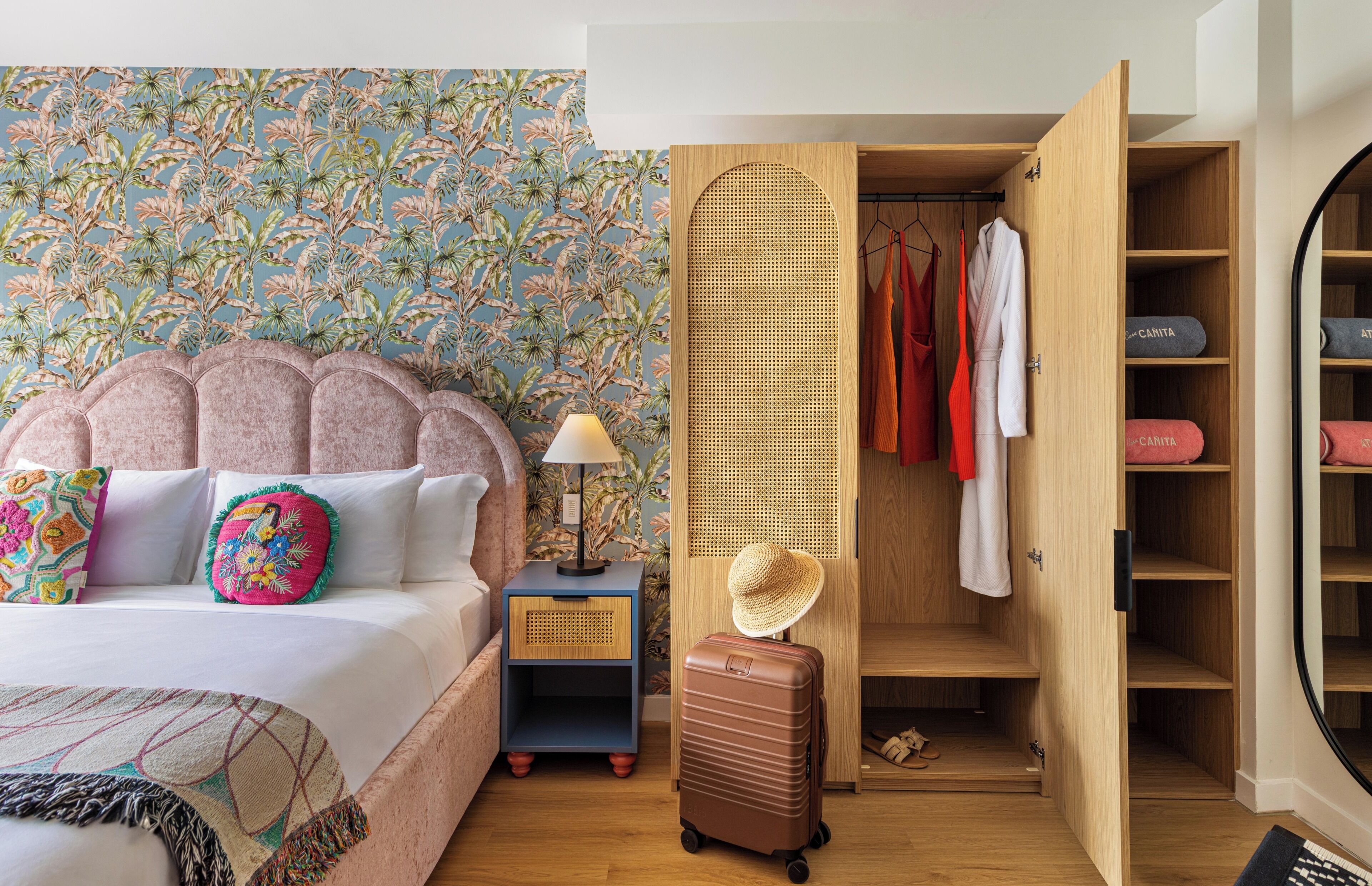 Ein Schlafzimmer mit Bett, Nachttisch, Koffer, Kleiderschrank und einer Wand mit tropischem Muster.