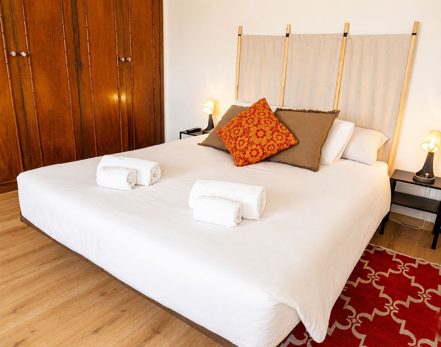 A neatly made bed with white linens and two rolled towels, a wooden wardrobe, a nightstand with a lamp, and a red patterned rug.