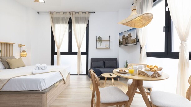 A modern hotel room with a bed, a sofa, a dining table with chairs, a wicker lamp, and a breakfast basket. - Travel Habitat Nou Mestalla 3 (Valencia)