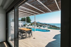 Ein Balkon mit Holztisch und -stühlen, ein Schwimmbad und Blick auf das Meer sowie entfernte Gebäude.