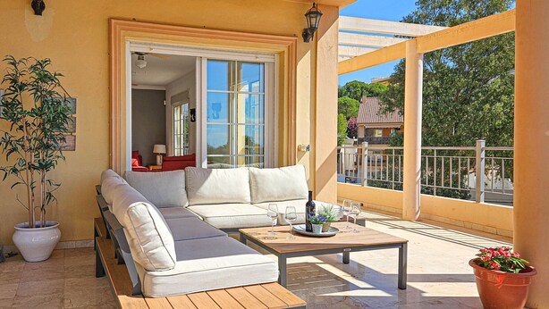 Un balcone illuminato dal sole con un divano bianco, un tavolino di legno e una pianta in vaso.