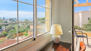 Una stanza con un'ampia finestra vista su una località costiera, un tavolo in legno con una lampada e un balcone con sedie.