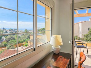 Une pièce dotée d’une grande fenêtre donnant sur une ville côtière, d’une table en bois avec une lampe et d’un balcon avec des chaises.