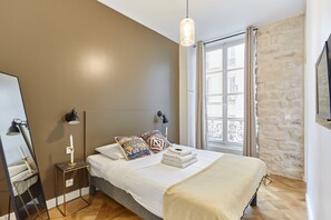 Une chambre moderne avec un grand lit, une table de chevet avec une lampe, un miroir et une fenêtre donnant sur des immeubles.