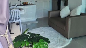 A modern living room with a brown sofa, a white coffee table, and a large green plant. A kitchen with white cabinets and a microwave is visible in the background. - Charming Condo in Wichit Comfort Meets Convenience (Wichit)