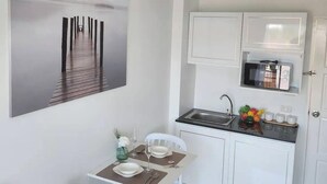 A compact kitchen with white cabinets, a microwave, and a sink. There is a dining area with a table and chairs, and a framed picture on the wall. - Charming Condo in Wichit Comfort Meets Convenience (Wichit)