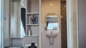 A hotel room with a bed, a bedside table with a lamp and a plant, a bathroom with a sink and mirror, and a shower area. - Charming Condo in Wichit Comfort Meets Convenience (Wichit)
