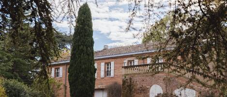 A brick building with a tall tree in front, surrounded by greenery.