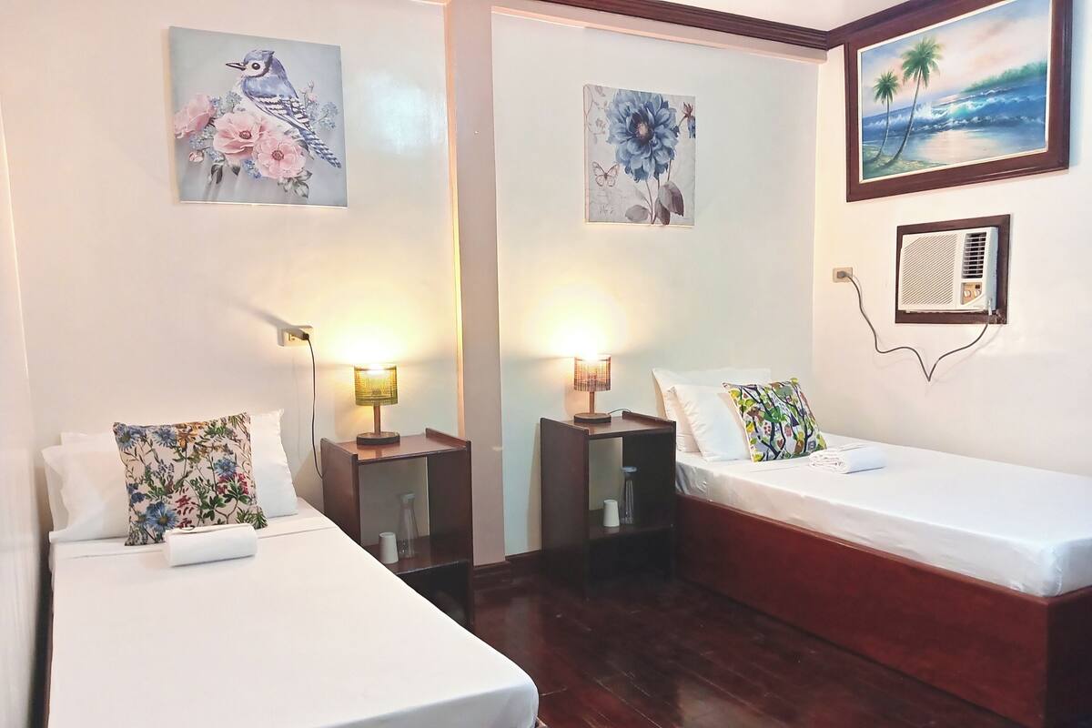 Ein Hotelzimmer mit zwei Einzelbetten, einem Nachttisch mit Lampe, einem kleinen Tisch und Wandaircondition.