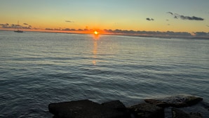 Atardecer sobre un mar tranquilo con un velero y costa rocosa.