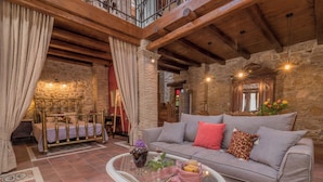 A cozy room with a stone wall, a wooden ceiling, a grey sofa with red and patterned cushions, a glass coffee table with flowers and candles, and a bed with a gold frame. - Authentic Zakynthos Town Villa * 3 open-plan bedrooms * Sleeps <7 * Local Stay (Zakinthos)
