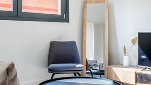 Ein modernes Zimmer mit einem großen Spiegel, einem grauen Sessel, einem Schreibtisch mit einem Flachbildfernseher und einem Fenster mit Blick auf ein Backsteingebäude.