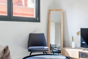 Ein modernes Zimmer mit einem großen Spiegel, einem grauen Sessel, einem Schreibtisch mit einem Flachbildfernseher und einem Fenster mit Blick auf ein Backsteingebäude.