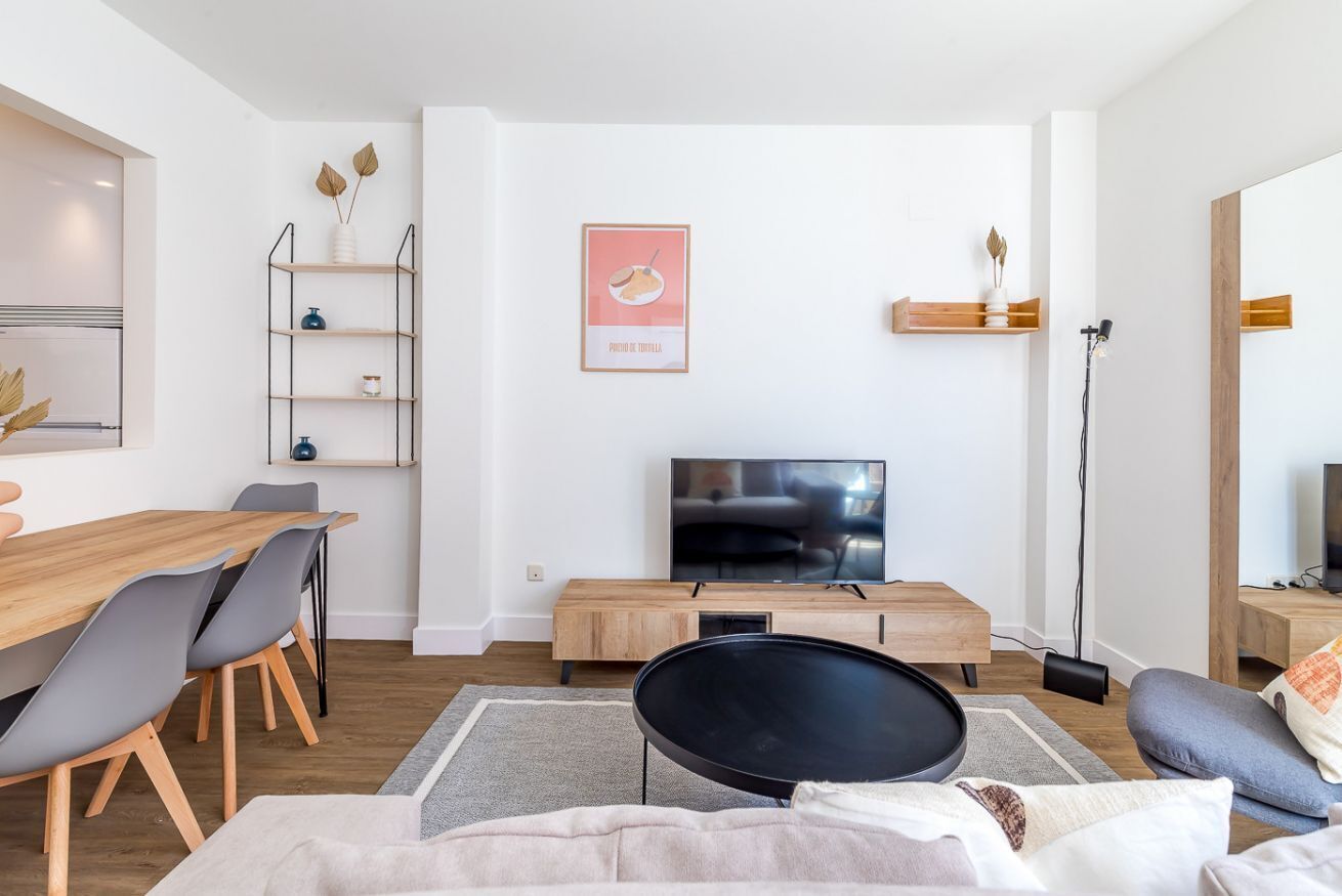 A modern living room with a wooden dining table, grey chairs, a black coffee table, a flat-screen TV, and a bookshelf.