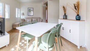 Ein heller Raum mit einem weißen Tisch und Stühlen, einem Sofa und einem Schrank mit Zierpflanzen.