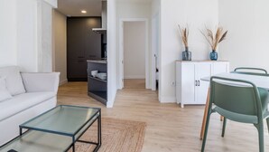 Ein modernes Wohnzimmer mit einer Couch, einem Glastisch und einem Essbereich mit Tisch und Stühlen. Im Hintergrund ist eine Küche mit Schränken und einer Arbeitsfläche zu sehen.