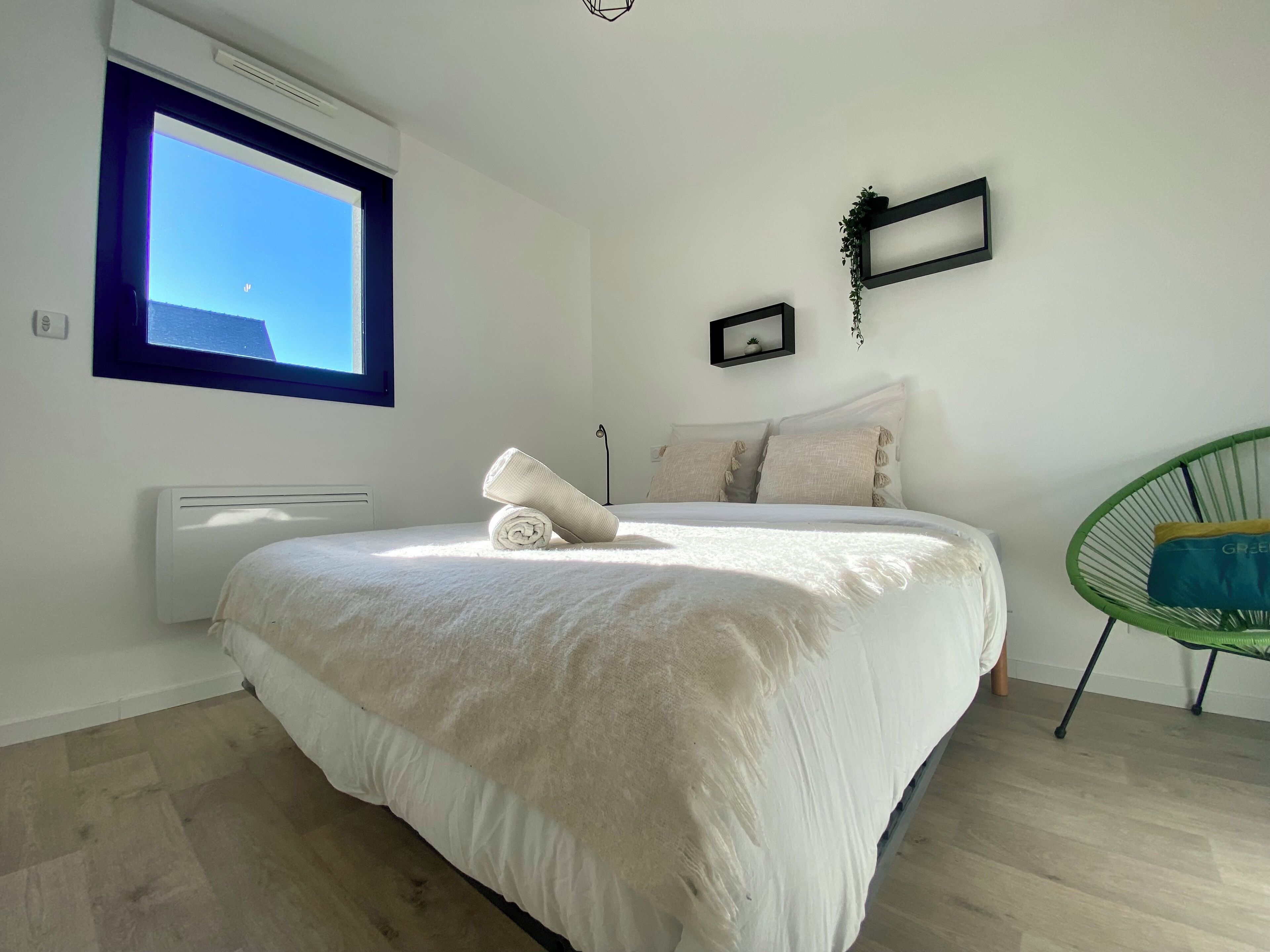 Een slaapkamer met een groot bed, een raam, een radiator, een groene stoel en een kleine plant aan de muur.