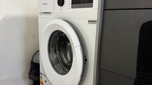 A front-loading washing machine with a control panel, located in a laundry room with a wooden floor and a grey door. - Le Relais de David - Limoges (Limoges)