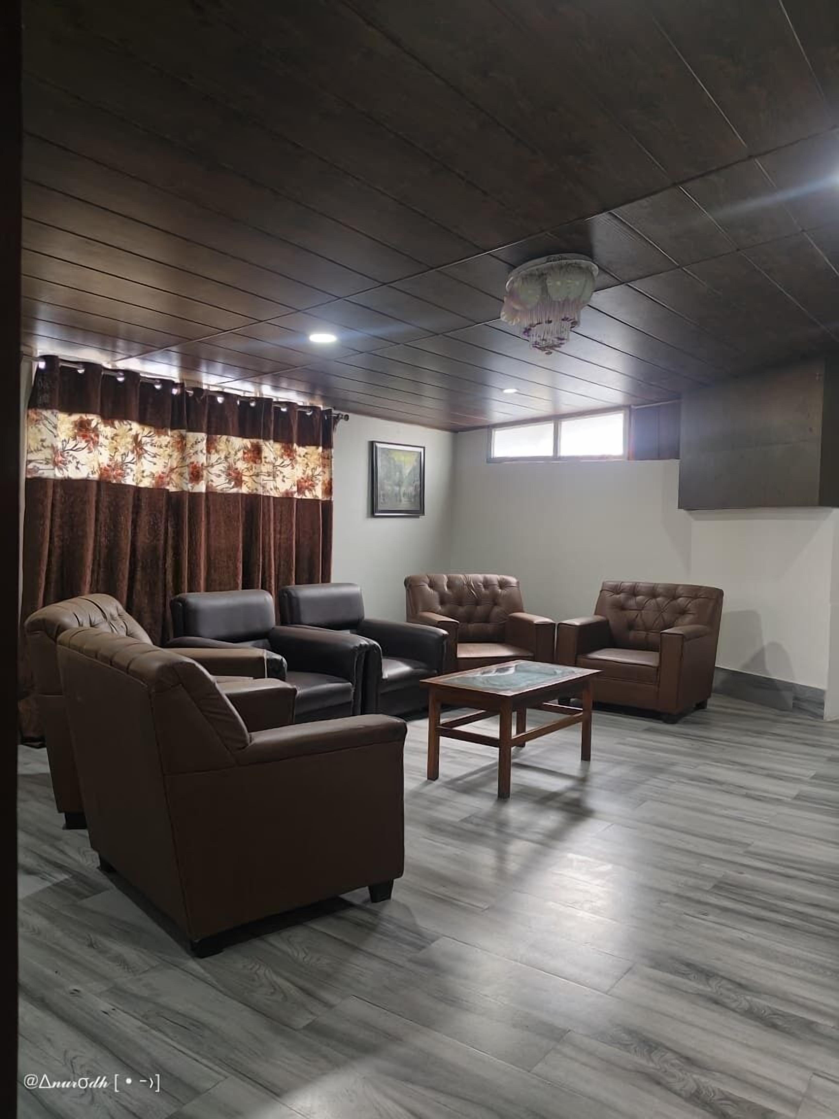 Ein Zimmer mit Holzbalkendecke, einem großen Fenster mit Vorhängen und einem gerahmten Bild an der Wand. Es gibt drei Ledersofas und einen Holztisch.