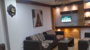 A living room with a sofa, coffee table, and TV. There is a clock on the wall and a decorative item on the left. - WORMWOOD SYNERGY NEW MANDALA  (Darjeeling)