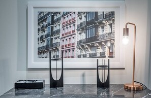 Una fotografía enmarco de un edificio con balcones, una lámpara de pie moderna y un caja de almacenamiento negra sobre una superficie de mármol.