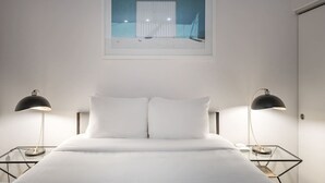 Une chambre à coucher avec un lit, deux lampes de chevet, une fenêtre donnant sur un immeuble et un tableau encadré au mur.