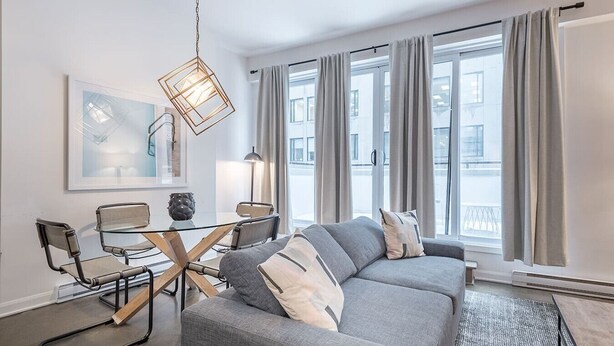Un salon moderne avec un canapé d’angle gris, une table à manger en verre et des chaises en métal, un luminaire suspendu et de grandes fenêtres avec des rideaux.