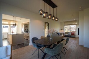 Una cocina moderna con una isla central, un comedor con mesa de madera oscura y sillas tapizadas en gris, y una sala de estar con sofá y televisor.