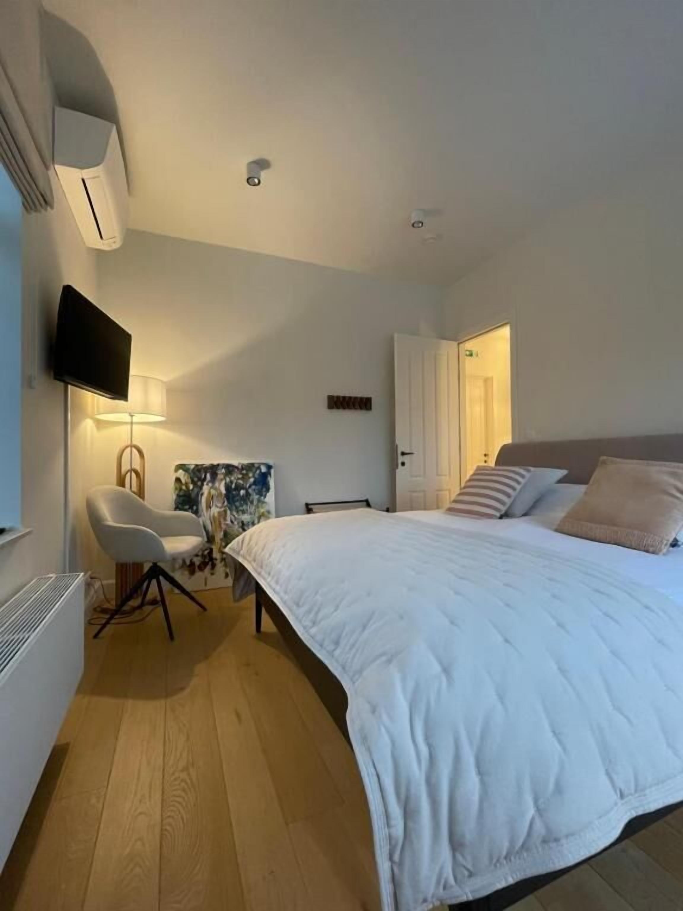 A modern bedroom with a large bed, a desk with a chair, a flat-screen TV, and a wall-mounted air conditioner.