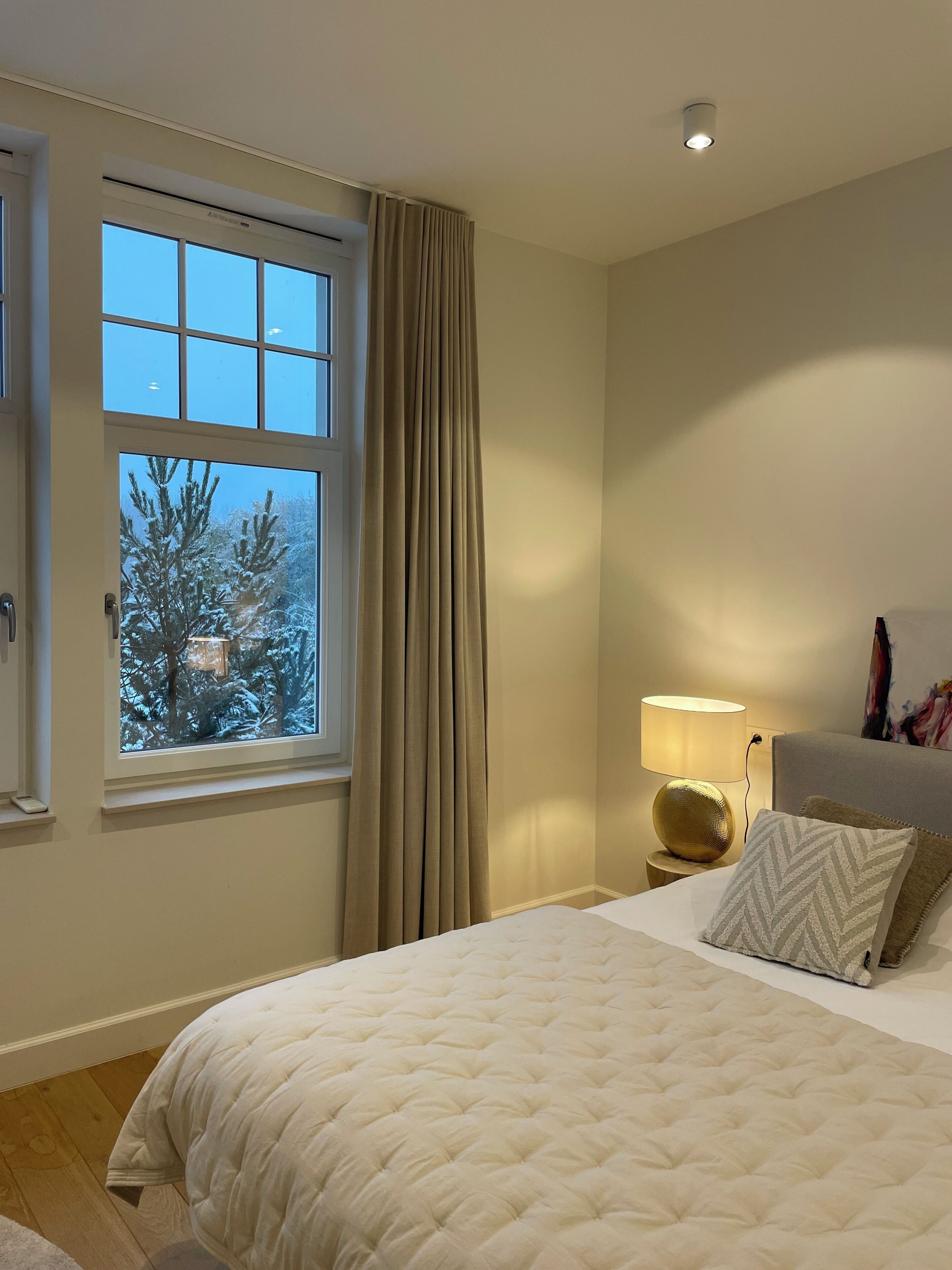 A bedroom with a large window, a bed with a white duvet, a bedside lamp, and a painting on the wall.