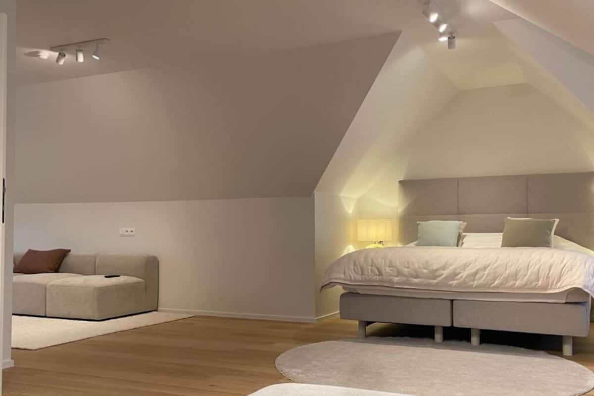 A bedroom with a sloped ceiling, a bed with a headboard, a bedside table, a lamp, and a sofa.