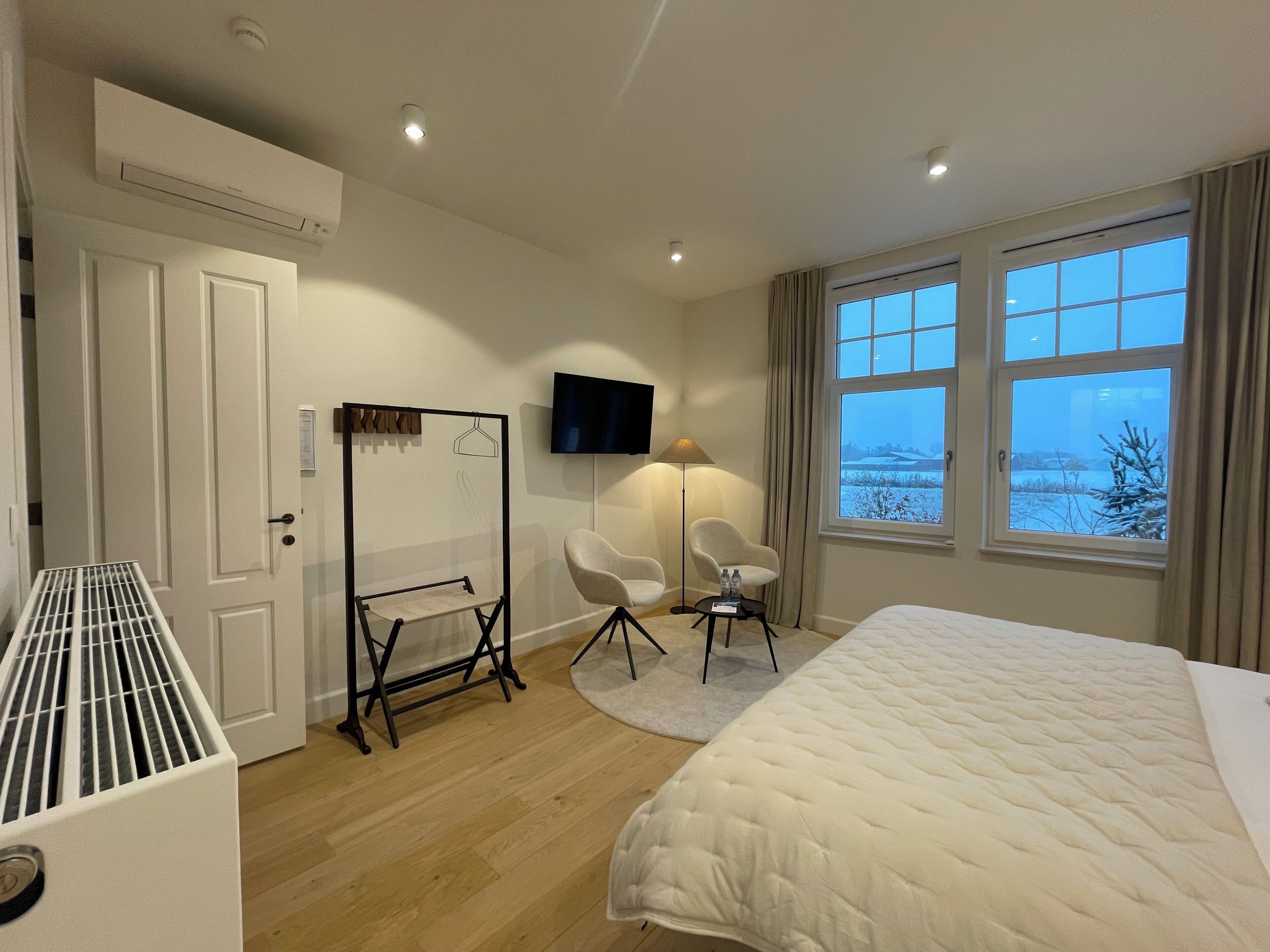 A hotel room with a bed, a seating area with two chairs, a flat-screen TV mounted on the wall, and a window with a view of a snowy landscape.