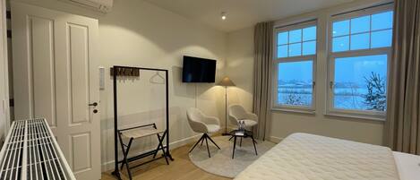 A hotel room with a bed, a seating area with two chairs, a flat-screen TV mounted on the wall, and a window with a view of a snowy landscape.