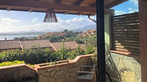 Un balcone con un muro in pietra, una sedia e vista su una località costiera con case e montagne sullo sfondo.