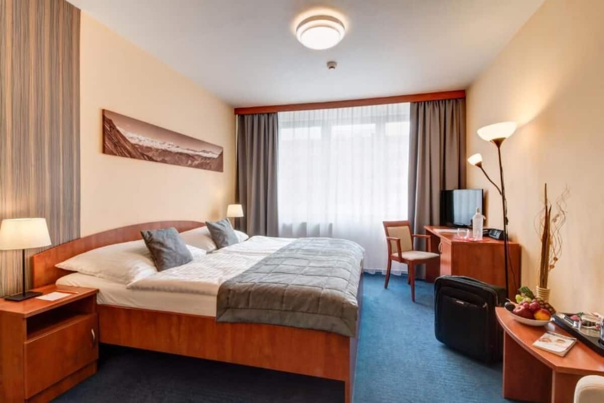 A hotel room with a bed, bedside table, desk, chair, and a desk lamp.