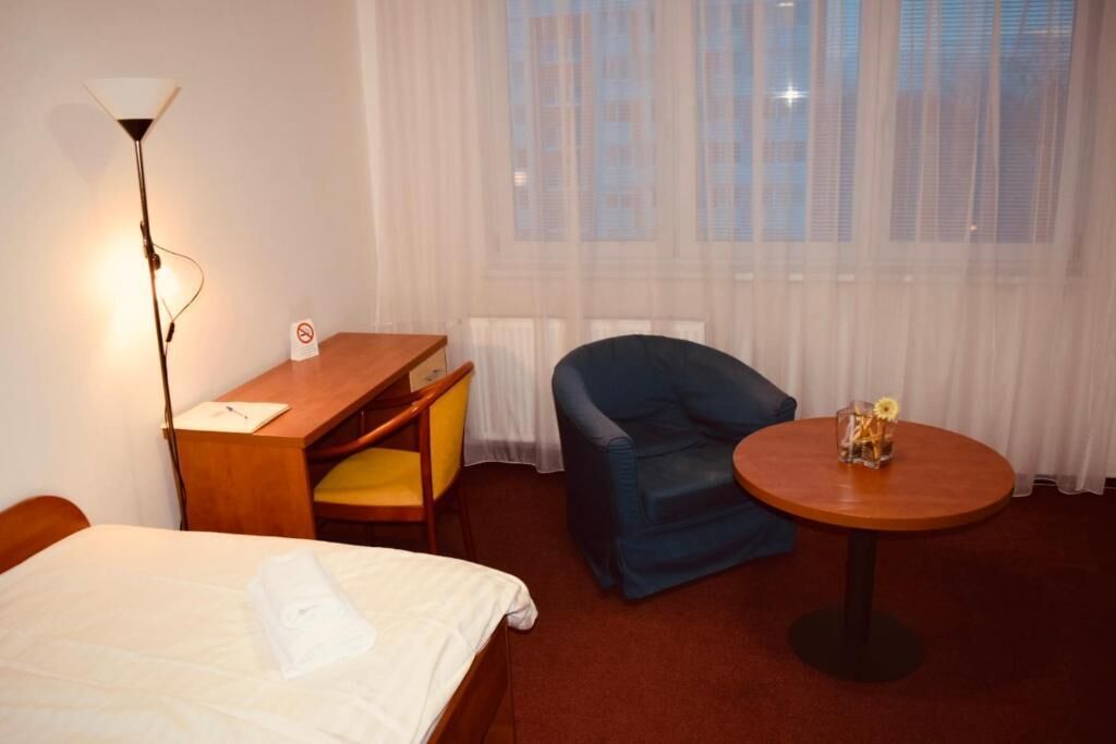 Habitación de hotel con cama, escritorio y silla, sillón azul, mesa redonda con un florero y ventana con cortinas traslúcidas.