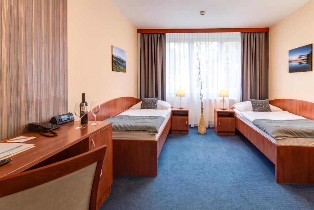 Ein Hotelzimmer mit zwei Einzelbetten, einem Schreibtisch mit Telefon und einem Weinglas sowie einem großen Fenster mit Vorhängen.
