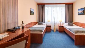 Ein Hotelzimmer mit zwei Einzelbetten, einem Schreibtisch mit Telefon und einem Weinglas sowie einem großen Fenster mit Vorhängen.