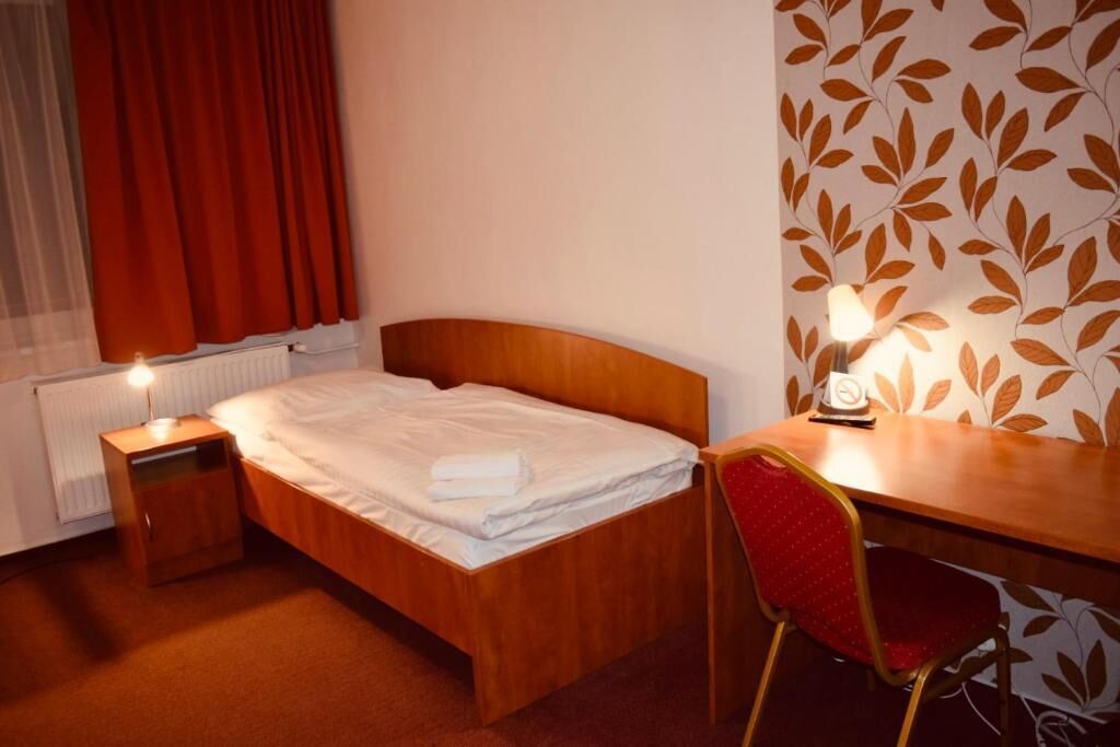 A hotel room with a wooden bed, a bedside table with a lamp, a red chair, and a wall with a leaf pattern.