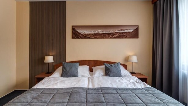 A hotel room with a large bed, two bedside tables with lamps, a painting on the wall, and a window with curtains. - LC Hotel Ostrava (Ostrava)